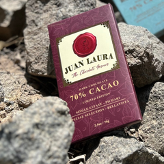 Juan Laura 70% Kakao Schokolade auf einem natürlichen, felsigen Hintergrund. Die burgunderrote Verpackung hebt sich kontrastreich von den Steinen ab und betont die Ursprünglichkeit und den natürlichen Ursprung des Kakaos.
