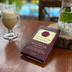 Juan Laura 70% Kakao Schokolade auf einem Holztisch neben einem Glas mit Milchkaffee und einer kleinen Pflanze in einer dekorativen Glasvase. Die Szene betont den Genussmoment mit der edlen, burgunderroten Verpackung der Schokolade.