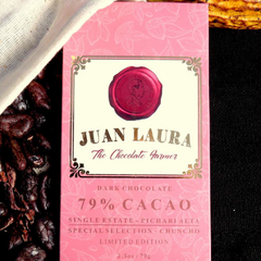 Verpackung der 79% dunklen Schokolade von Juan Laura mit rotem Siegel und rosa Muster, liegend auf einer schwarzen Oberfläche und teilweise bedeckt von weißem Stoff, umgeben von Kakaobohnen. Text: '79% CACAO', 'Single Estate - Pichari Alta', 'Special Selection - Chuncho', 'Limited Edition', '2.5 oz / 70 g'.