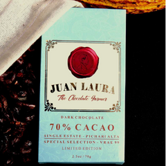 Verpackung der 70% dunklen Schokolade von Juan Laura mit rotem Siegel und hellblauem Muster, liegend auf einer schwarzen Oberfläche und teilweise bedeckt von weißem Stoff, umgeben von Kakaobohnen. Text: '70% CACAO', 'Single Estate - Pichari Alta', 'Special Selection - VRAE 99', 'Limited Edition', '2.5 oz / 70 g'.