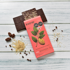 Kuyay Schokolade mit Quinoa, 70% Kakao, auf einem hellen Holzuntergrund. Die Schokoladentafel ist geöffnet und zeigt Quinoa-Stückchen in der Schokolade, umgeben von rohen Kakaobohnen, Quinoa und Kakaonibs als Dekoration.