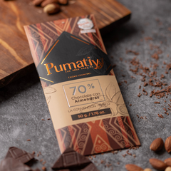 Eine Tafel Pumatiy Bitterschokolade mit 70% Kakaoanteil und Mandeln liegt auf einer grauen Oberfläche, umgeben von Kakaonibs und Mandeln. Die Verpackung hat ein rotes und braunes Muster und zeigt die Auszeichnung der Peru Chocolate Awards 2022.