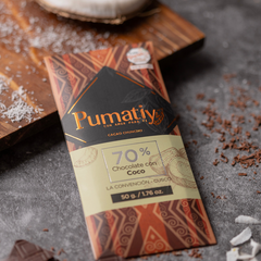 Eine Tafel Pumatiy Bitterschokolade mit 70% Kakaoanteil und Kokosnuss liegt auf einer grauen Oberfläche, umgeben von Kakaonibs und Kokosraspeln. Die Verpackung hat ein rotes und braunes Muster und zeigt die Auszeichnung der Peru Chocolate Awards 2022.