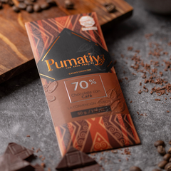 Eine Tafel Pumatiy Bitterschokolade mit 70% Kakaoanteil und Kaffee liegt auf einer grauen Oberfläche, umgeben von Kakaonibs und Kaffeebohnen. Die Verpackung hat ein rotes und braunes Muster und zeigt die Auszeichnung der Peru Chocolate Awards 2022.