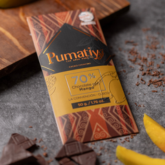 Eine Tafel Pumatiy Bitterschokolade mit 70% Kakaoanteil und Mango liegt auf einer grauen Oberfläche, umgeben von Kakaonibs und Mangostücken. Die Verpackung hat ein rotes und braunes Muster und zeigt die Auszeichnung der Peru Chocolate Awards 2022.