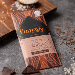 Eine Tafel Pumatiy Bitterschokolade mit 70% Kakaoanteil und Maras-Salz liegt auf einer grauen Oberfläche, umgeben von Kakaonibs und Salzflocken. Die Verpackung hat ein rotes und braunes Muster und zeigt die Auszeichnung der Peru Chocolate Awards 2022.
