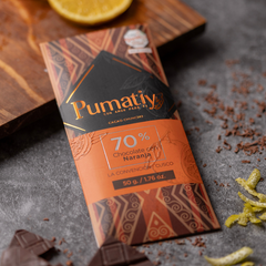 Eine Tafel Pumatiy Bitterschokolade mit 70% Kakaoanteil und Orange liegt auf einer grauen Oberfläche, umgeben von Kakaonibs und Orangenschalen. Die Verpackung hat ein rotes und braunes Muster und zeigt die Auszeichnung der Peru Chocolate Awards 2022.
