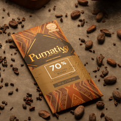 Eine Tafel Pumatiy Bitterschokolade mit 70% Kakaoanteil liegt auf einer grauen Oberfläche, umgeben von Kakaobohnen und Kakaonibs. Die Szene vermittelt eine rustikale und handwerkliche Atmosphäre.
