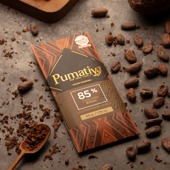 Eine Tafel Pumatiy Bitterschokolade mit 85% Kakaoanteil liegt auf einer grauen Oberfläche, umgeben von Kakaobohnen und Kakaonibs. Ein Holzlöffel mit zerkleinertem Kakao ist ebenfalls im Bild zu sehen. Die Verpackung zeigt die Auszeichnung der Peru Chocolate Awards 2022 und vermittelt eine rustikale und handwerkliche Atmosphäre.