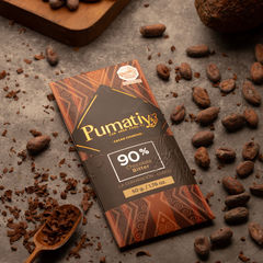Eine Tafel Pumatiy Bitterschokolade mit 90% Kakaoanteil liegt auf einer grauen Oberfläche, umgeben von Kakaobohnen und Kakaonibs. Ein Holzlöffel mit zerkleinertem Kakao ist ebenfalls im Bild zu sehen. Die Szene vermittelt eine rustikale und handwerkliche Atmosphäre.