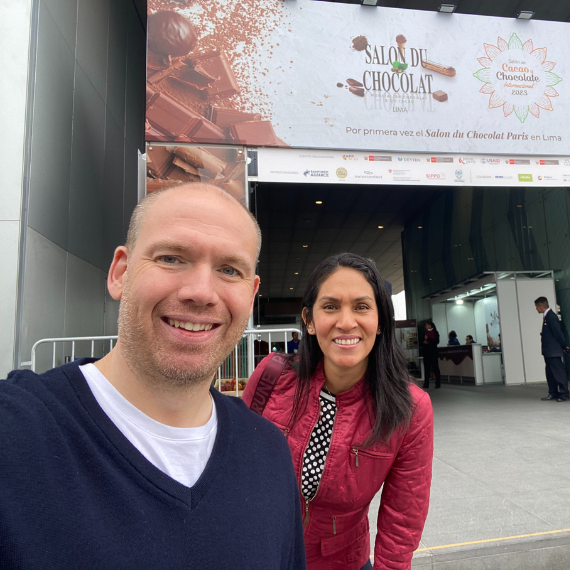 Ein Mann und eine Frau stehen vor dem Eingang zu einer Veranstaltung mit dem Banner 'Salon du Chocolat 2023'. Beide lächeln in die Kamera. Der Mann trägt ein dunkelblaues Oberteil und die Frau eine rote Jacke.