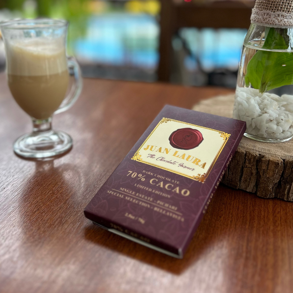 Juan Laura 70% Kakao Schokolade auf einem Holztisch neben einem Glas mit Milchkaffee und einer kleinen Pflanze in einer dekorativen Glasvase. Die Szene betont den Genussmoment mit der edlen, burgunderroten Verpackung der Schokolade.