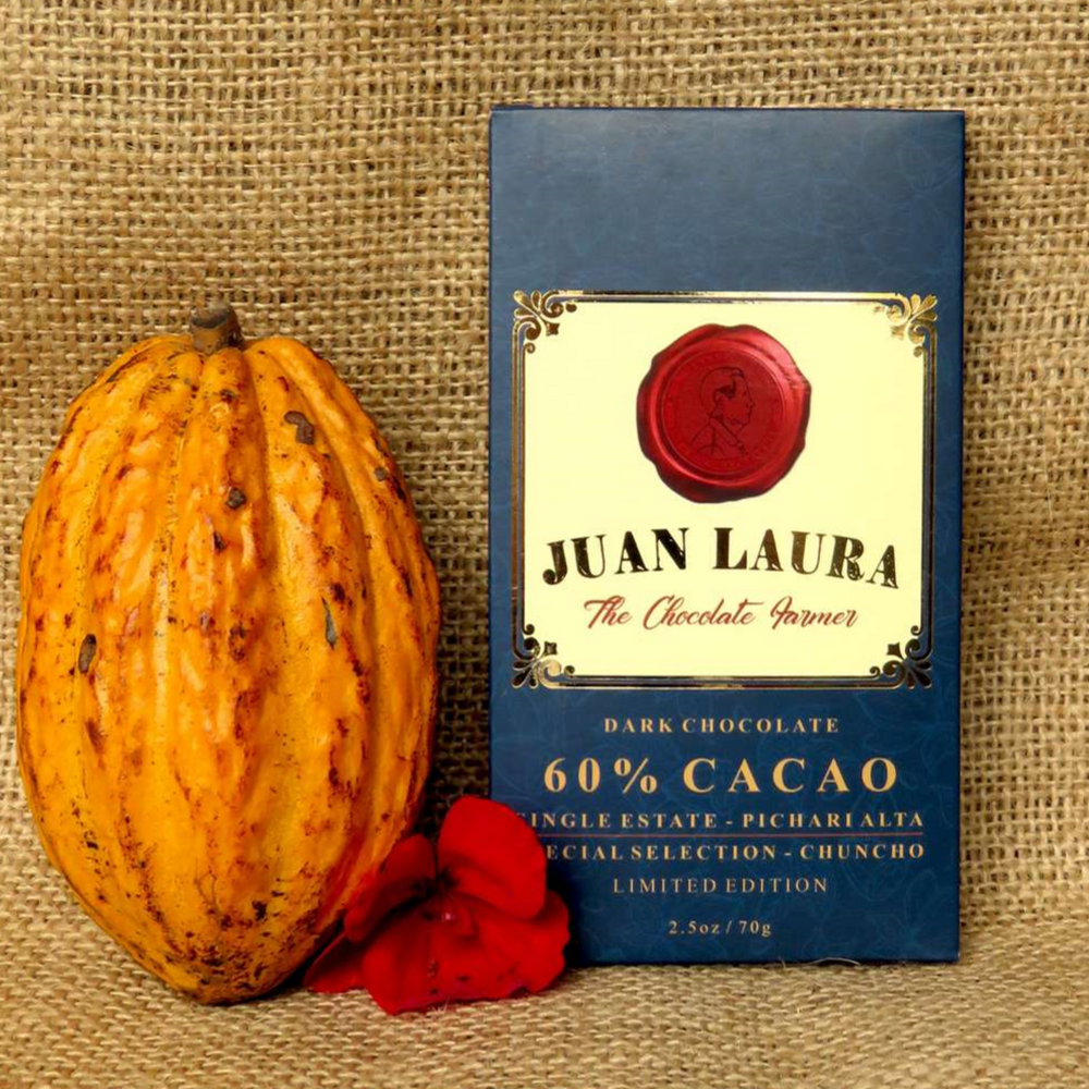 Verpackung der dunklen Schokolade mit 60% Kakaoanteil von Juan Laura - The Chocolate Farmer. Die Verpackung ist dunkelblau mit einem roten Siegel und goldenen Verzierungen. Daneben liegt eine große, gelb-orangefarbene Kakaofrucht und ein rotes Blütenblatt, alles vor einem Hintergrund aus Jutestoff. Auf der Verpackung steht '60% CACAO', 'Single Estate - Pichari Alta', 'Special Selection - Chuncho', 'Limited Edition' und '2.5 oz / 70 g'.