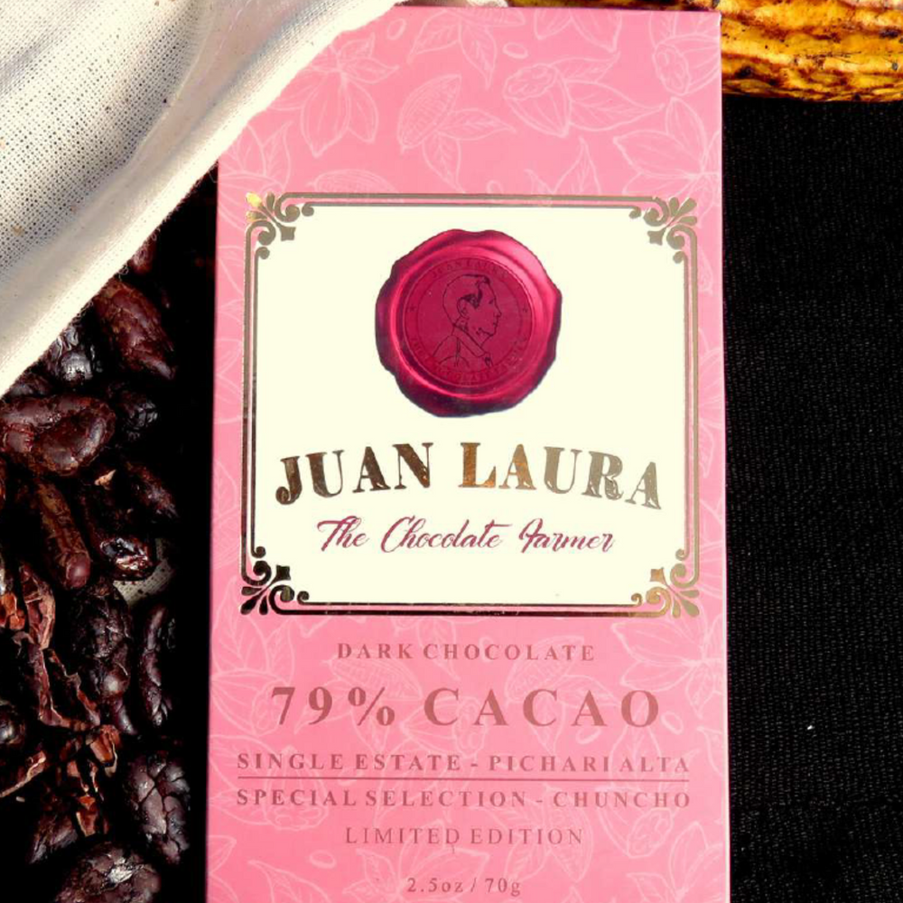 Verpackung der 79% dunklen Schokolade von Juan Laura mit rotem Siegel und rosa Muster, liegend auf einer schwarzen Oberfläche und teilweise bedeckt von weißem Stoff, umgeben von Kakaobohnen. Text: '79% CACAO', 'Single Estate - Pichari Alta', 'Special Selection - Chuncho', 'Limited Edition', '2.5 oz / 70 g'.