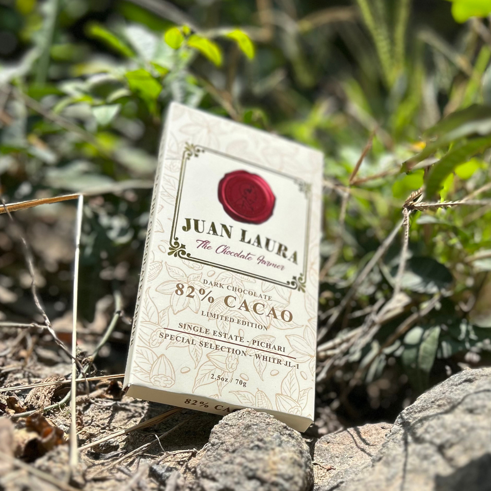 Juan Laura 82% Kakao Schokolade auf einem natürlichen, waldähnlichen Hintergrund platziert, umgeben von grünen Pflanzen und Felsen. Die cremefarbene Verpackung hebt die Ursprünglichkeit und handwerkliche Qualität der Single-Estate-Schokolade hervor.