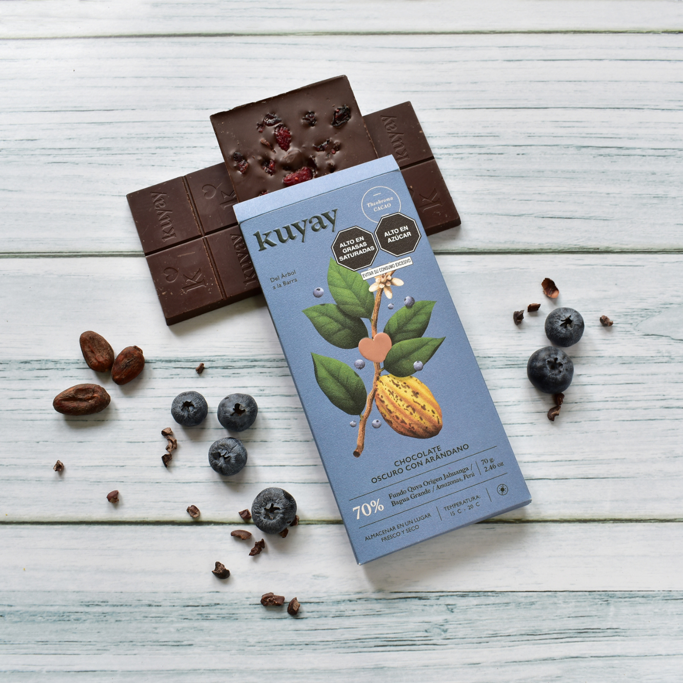 Kuyay 70% Dunkle Schokolade mit Blaubeeren, neben der geöffneten Schokoladentafel, Blaubeeren und Kakaobohnen auf einem weißen Holzuntergrund präsentiert. Die Schokoladentafel zeigt auf der Oberfläche eingestreute getrocknete Blaubeeren und Kakaonibs.