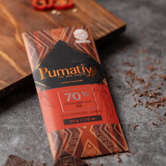 Eine Tafel Pumatiy Bitterschokolade mit 70% Kakaoanteil und peruanischem Chili liegt auf einer grauen Oberfläche, umgeben von Kakaonibs und Chilistücken. Die Verpackung hat ein rotes und braunes Muster und zeigt die Auszeichnung der Peru Chocolate Awards 2022.
