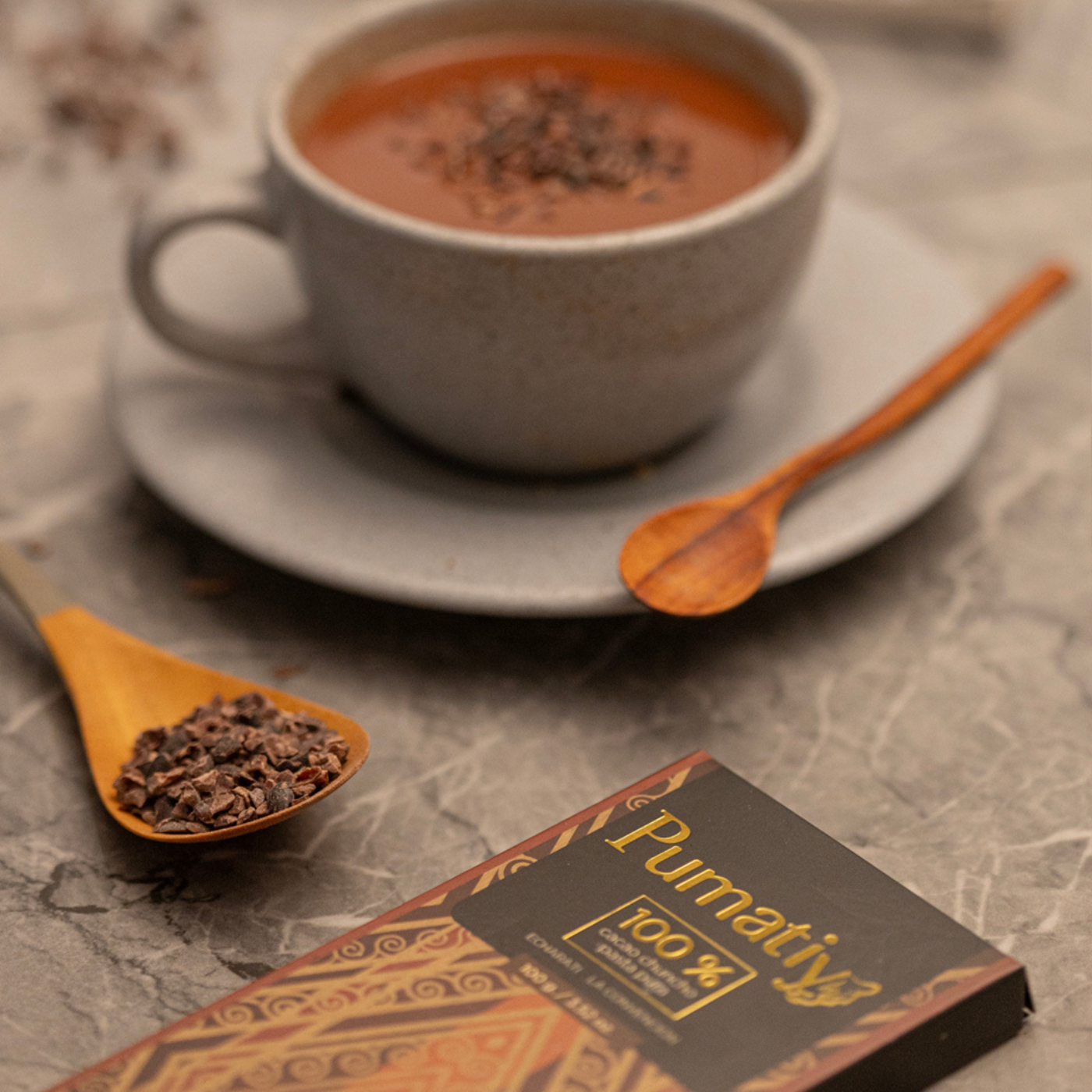 Eine Tasse heiße Schokolade, zubereitet mit Pumatíy 100 % Cacao Chuncho Pasta Pura, auf einer Untertasse mit einem Holzlöffel. Im Vordergrund liegt eine Pumatíy-Schokoladentafel und ein Holzlöffel, gefüllt mit Kakaonibs. Die Szene ist auf einer Marmoroberfläche arrangiert.